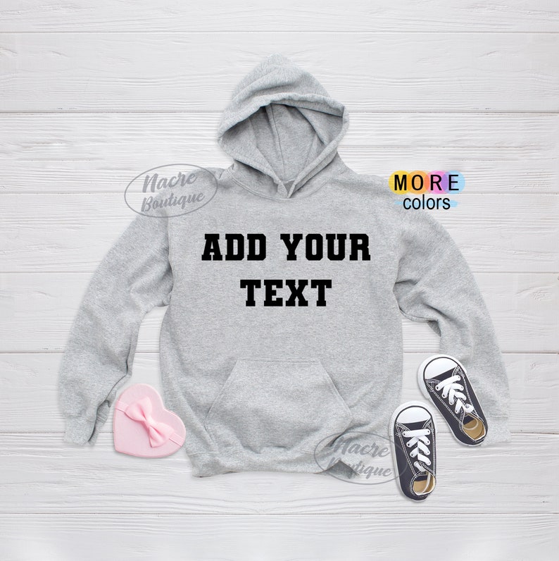 May include: A light gray hoodie with a front pocket and hood. The text "ADD YOUR TEXT" is printed in black block letters across the chest. A pink heart-shaped box and a pair of black and white sneakers are next to the hoodie.