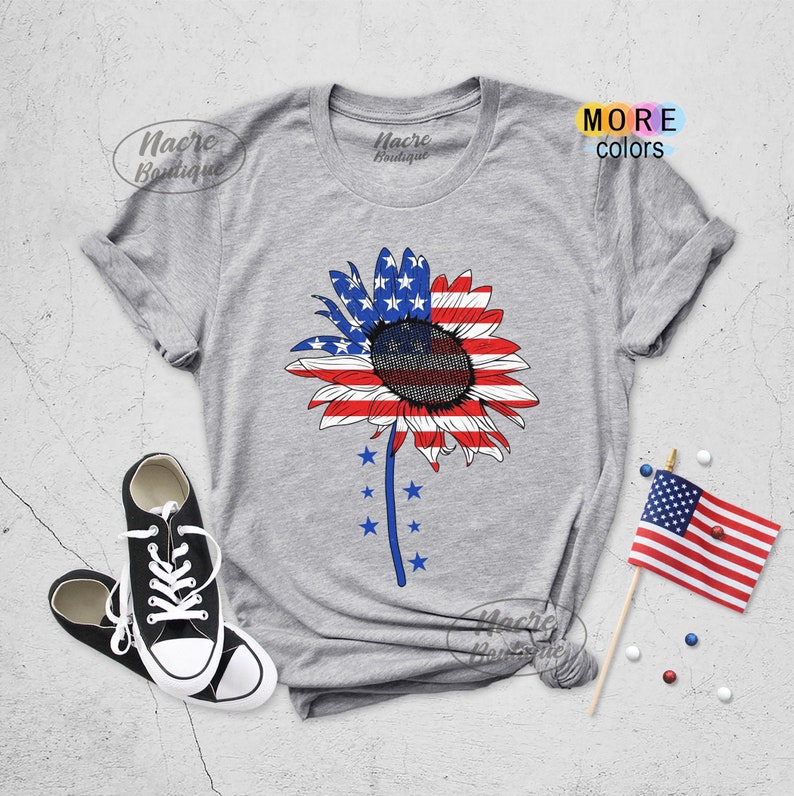 May include: A gray t-shirt with a sunflower design featuring the American flag colors. The sunflower has blue stars around the stem. The shirt is on a gray surface with a pair of black sneakers and a small American flag.