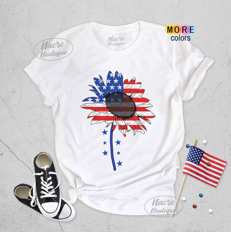 May include: White t-shirt with a sunflower design featuring the American flag colors of red, white, and blue. The sunflower has a dark brown center and is surrounded by white stars.