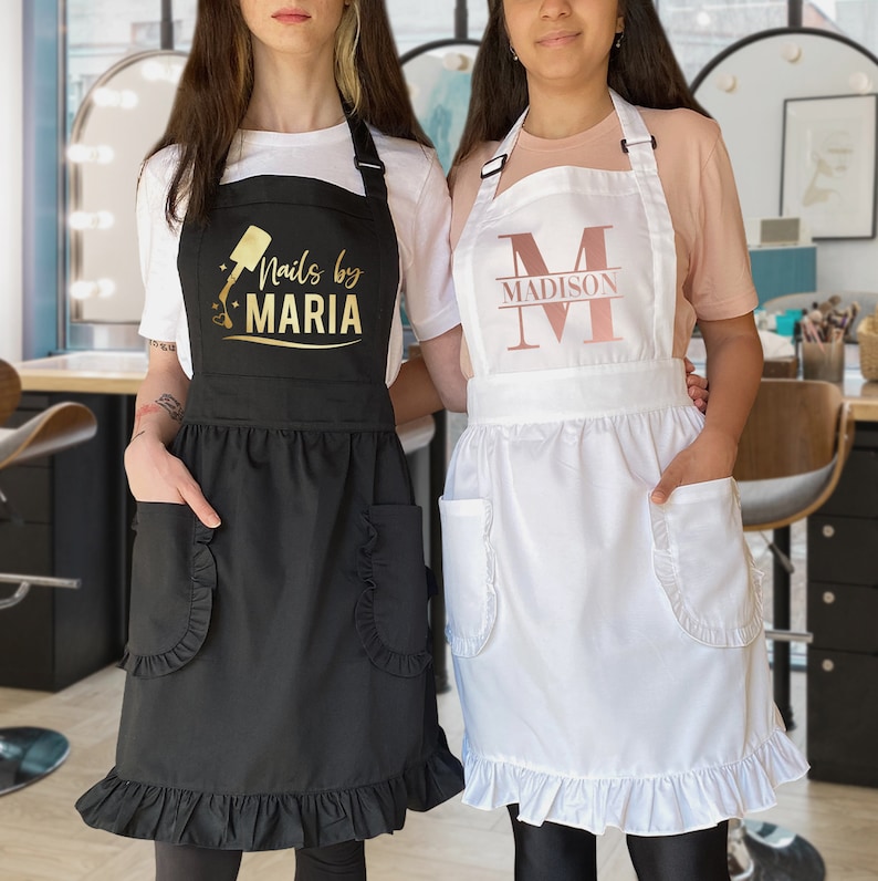 Puede incluir: Dos delantales negros y blancos con bordes con volantes. Un delantal tiene el texto "Nails by Maria" en letras doradas y un gr&aacute;fico de botella de esmalte de u&ntilde;as. El otro delantal tiene una gran "M" dorada con el texto "Madison" debajo.