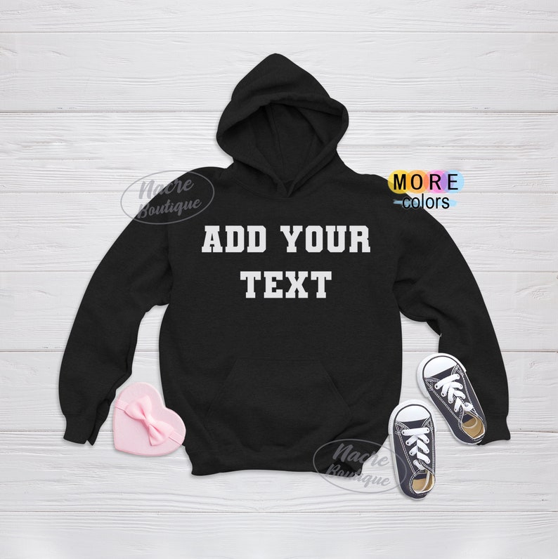 May include: Black hooded sweatshirt with the text "ADD YOUR TEXT" in white block letters. The sweatshirt has a front pocket and a hood. A pink heart-shaped box and a pair of small black sneakers are also in the image.