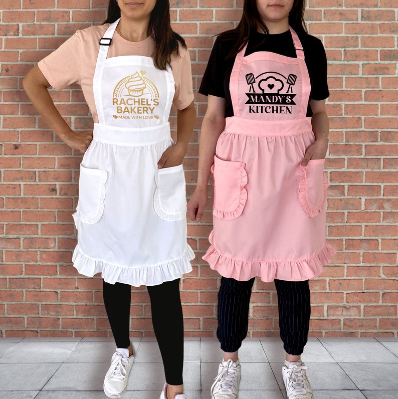 Puede incluir: Dos mujeres usando delantales con texto personalizado. El delantal de la izquierda es blanco con texto dorado que dice "Rachel's Bakery Made with Love". El delantal de la derecha es rosa con texto negro que dice "Mandy's Kitchen".