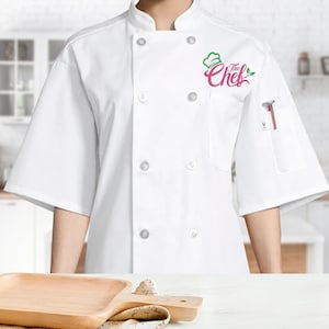 May include: A white double-breasted chef coat with short sleeves and a mandarin collar. The front features white buttons and an embroidered logo on the left chest that reads The Chef in pink script with a green chef hat and leaf icon above it.
