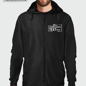May include: A black zip-up hooded windbreaker jacket featuring a white embroidered rectangular graphic on the left chest. The graphic contains the text Your EMBROIDERY TEXT here. The jacket has long sleeves, a hood with drawstrings, and a front zipper.