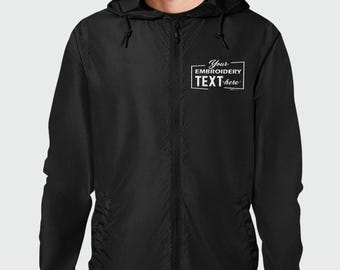 May include: A black zip-up hooded windbreaker jacket featuring a white embroidered rectangular graphic on the left chest. The graphic contains the text Your EMBROIDERY TEXT here. The jacket has long sleeves, a hood with drawstrings, and a front zipper.