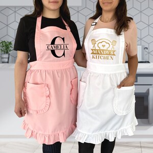 Puede incluir: Dos delantales rosa y blanco con bordes con volantes. Un delantal tiene el nombre "Camelia" en letras negras y el otro delantal tiene el texto "Mandy's Kitchen" en letras doradas.