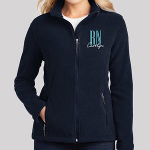 May include: A navy blue full-zip fleece jacket featuring a high collar and two side pockets. The left chest is embroidered with the letters RN in a light blue sans-serif font, positioned above the name Carolyn in a white cursive script font.