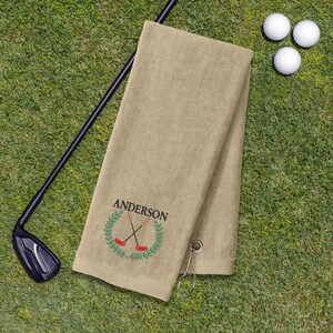 a tan towel with a personalized name embroidered on it, along with a golf club and three golf balls on a grassy surface.