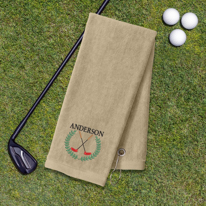 a tan towel with a personalized name embroidered on it, along with a golf club and three golf balls on a grassy surface.