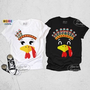 May include: Two short-sleeved t-shirts, one white and one black, featuring turkey face designs. The turkey faces have yellow beaks, red wattles, and colorful feather headdresses. The white shirt has a turkey face with eyelashes. The black shirt has a turkey face with a different feather headdress.