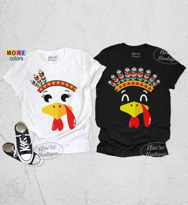 May include: Two short-sleeved t-shirts, one white and one black, featuring turkey face designs. The turkey faces have yellow beaks, red wattles, and colorful feather headdresses. The white shirt has a turkey face with eyelashes. The black shirt has a turkey face with a different feather headdress.