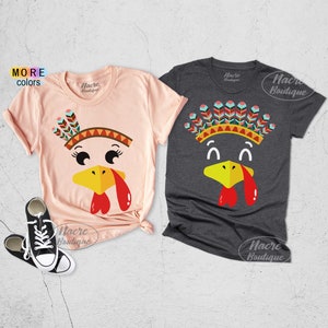 May include: Two short-sleeved t-shirts with turkey face designs. One is light pink, the other is dark gray. Each shirt features a turkey face with a yellow beak, red wattle, and a Native American headdress. The text "More Colors" is visible.