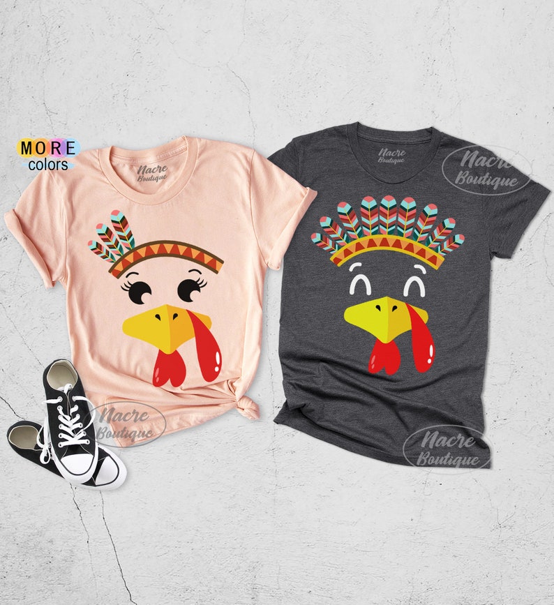 May include: Two short-sleeved t-shirts with turkey face designs. One is light pink, the other is dark gray. Each shirt features a turkey face with a yellow beak, red wattle, and a Native American headdress. The text "More Colors" is visible.