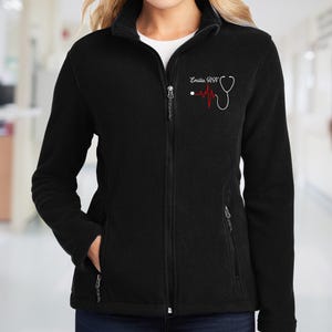 May include: A black fleece zip-up jacket featuring a white embroidered stethoscope design on the left chest. The embroidery includes a red heartbeat line and the text "Emilia RN" in a white script font. The jacket has a high collar and pockets.
