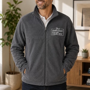 May include: A dark charcoal gray fleece jacket with a full-length front zipper, a high collar, and long sleeves with elastic cuffs. The left chest features a rectangular embroidered design with the text Your EMBROIDERY TEXT here.