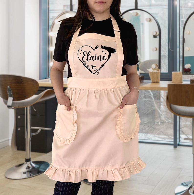 Puede incluir: Un delantal rosa con un dise&ntilde;o de coraz&oacute;n que presenta herramientas de peluquer&iacute;a y el nombre "Elaine". El delantal tiene dos bolsillos y un dobladillo con volantes.
