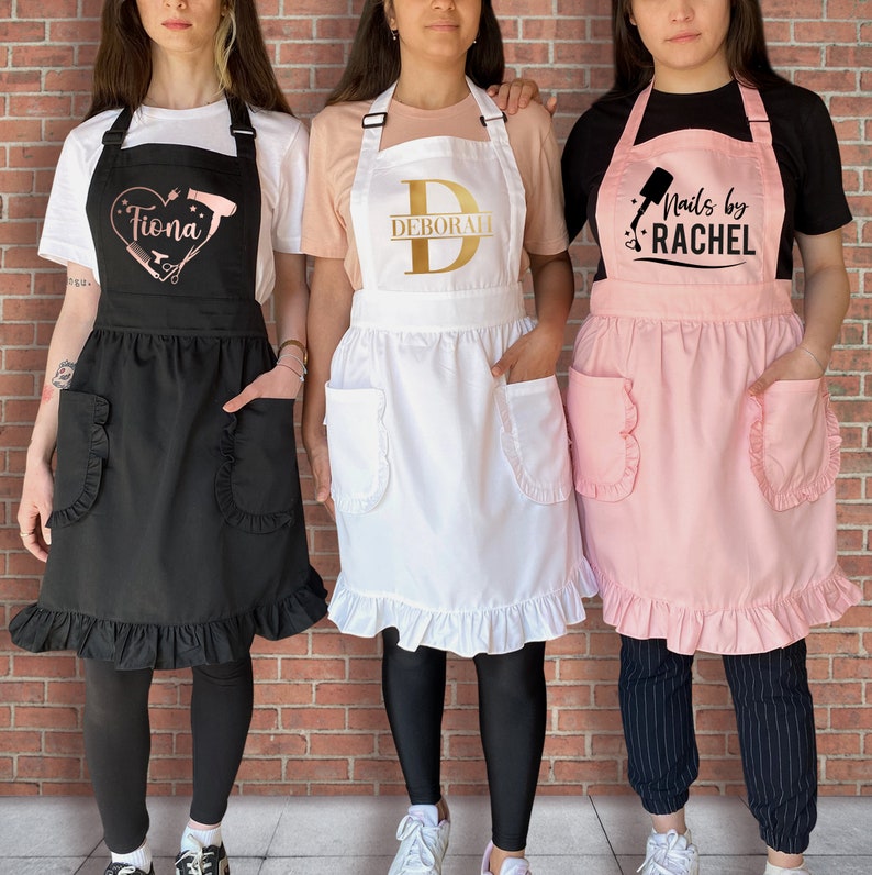 Puede incluir: Tres mujeres con diferentes delantales con texto personalizado. Los delantales son negro, blanco y rosa con bordes con volantes. El texto en los delantales dice "Tiona", "Deborah" y "Nails by Rachel".