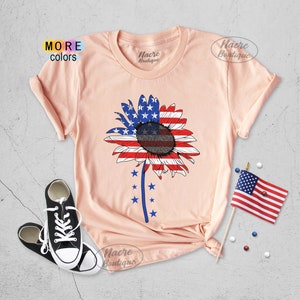 May include: A peach-colored t-shirt with a sunflower design. The sunflower is made up of the American flag colors, red, white, and blue. The shirt has the text "Nacre Boutique" on the front.