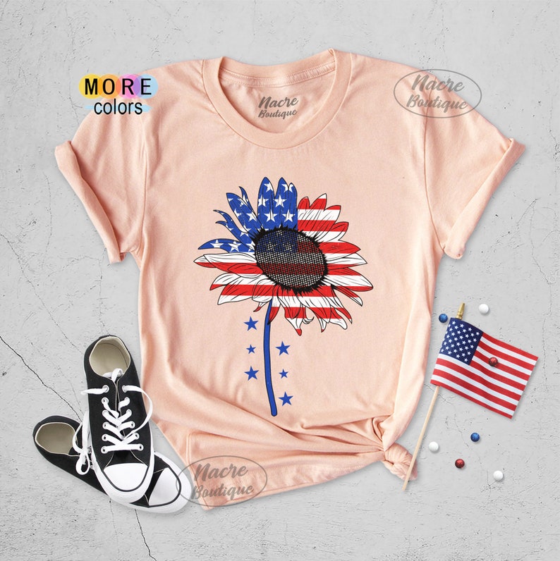 May include: A peach-colored t-shirt with a sunflower design. The sunflower is made up of the American flag colors, red, white, and blue. The shirt has the text "Nacre Boutique" on the front.