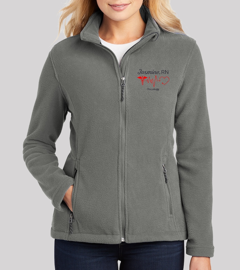 May include: Gray fleece jacket with a zipper closure and two pockets. The jacket has an embroidered design that says "Jasmine, RN" with a red heart and a medical symbol.