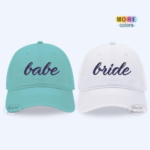 May include: Two baseball caps, one teal and one white. The teal cap has the word "babe" in cursive, and the white cap has the word "bride" in cursive. The caps have a curved brim and a fabric strap closure.