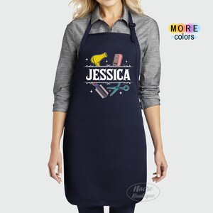 Puede incluir: Un delantal azul marino con un bolsillo blanco y un dise&ntilde;o con un secador de pelo, un peine, unas tijeras y el nombre "JESSICA".