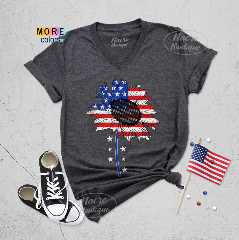 May include: A dark grey V-neck t-shirt with a sunflower design. The sunflower is made up of the American flag colors, red, white, and blue. The shirt has the text "Nacre Boutique" on the front.