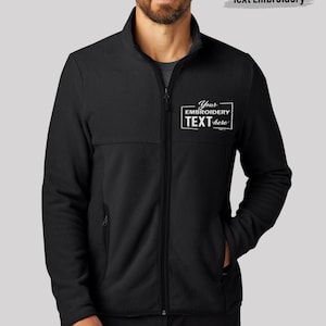 May include: A black fleece jacket with a full-length front zipper, a high collar, and long sleeves. The left chest features a white rectangular graphic with the text "Your EMBROIDERY TEXT here" printed in a mix of bold and script fonts.