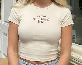 以下が含まれることがあります： 胸元に「your text embroidered here」という茶色の刺繍が施された、クリーム色の半袖リブTシャツ。クルーネックで体にフィットするシルエットで、ライトブルーのデニムジーンズと組み合わされています。