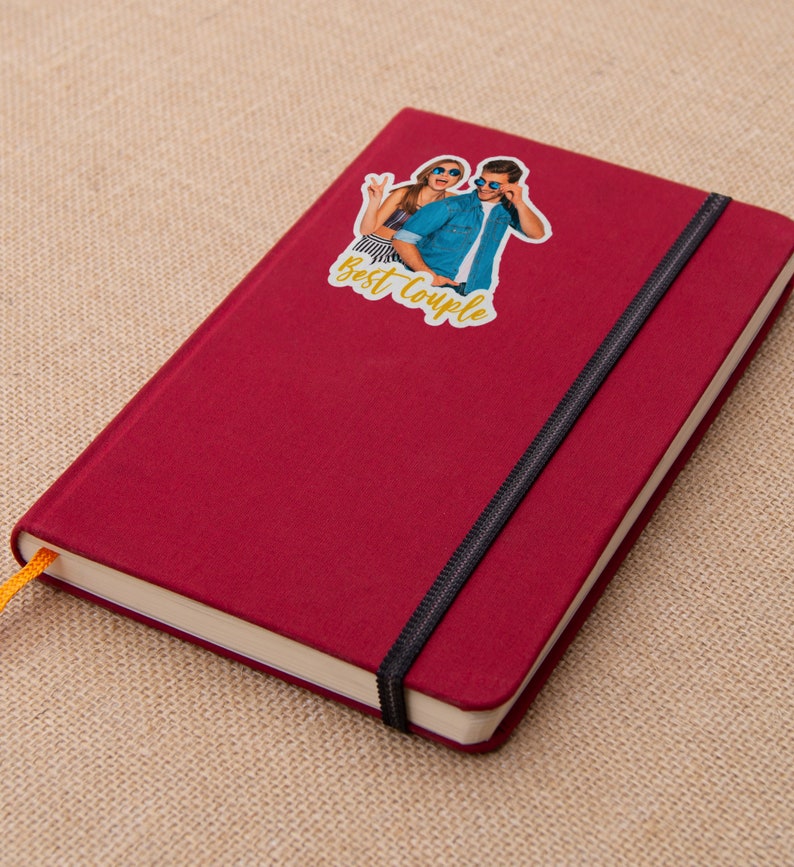 Puede incluir: Una libreta roja con cierre de banda el&aacute;stica negra. Una pegatina con una foto de una pareja y el texto "Best Couple" est&aacute; en la portada.