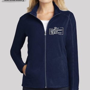 May include: A navy blue fleece jacket with a full-length front zipper and a high collar. The left chest features white embroidered text inside a rectangular frame that reads: Your EMBROIDERY TEXT here. The jacket has long sleeves and side pockets.