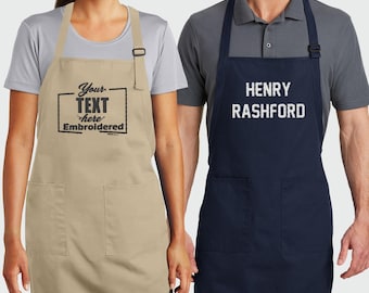 May include: A beige apron with black embroidered text reading Your TEXT here Embroidered and a navy blue apron with white embroidered text reading HENRY RASHFORD. Both aprons feature two front pockets, adjustable neck straps, and waist ties.