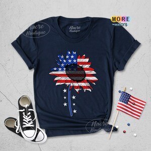 May include: A navy blue t-shirt with a sunflower design featuring the American flag colors. The sunflower has a black center and red, white, and blue petals. The shirt has a crew neck and short sleeves.