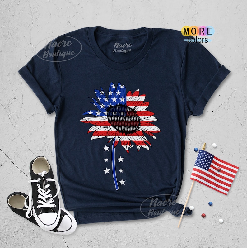 May include: A navy blue t-shirt with a sunflower design featuring the American flag colors. The sunflower has a black center and red, white, and blue petals. The shirt has a crew neck and short sleeves.