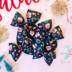 May include: Several navy blue fabric bows with a Christmas-themed pattern of gifts, wreaths, and candy canes are arranged on a pink surface. The word "falala" is written in red script above the bows. Decorative stars and small Christmas trees are also present.