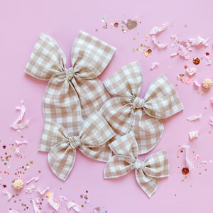 May include: Four beige and white gingham fabric bows of varying sizes are arranged on a pink surface. The bows are tied in the center and have a classic bow shape. Decorative confetti and small pumpkins are scattered around the bows.