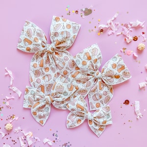 May include: Several fabric hair bows in varying sizes, featuring a repeating pattern of baked goods like pie slices, cupcakes, and pumpkins. The bows are cream-colored with orange, pink, and brown illustrations, set against a light pink background with confetti.
