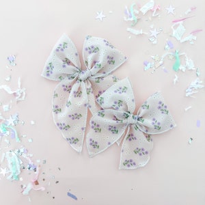 May include: Two floral hair bows with a light pink background. The bows are made of a light beige fabric with a small purple floral pattern. The bows are tied in a knot in the center.