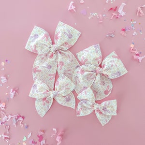 May include: Five fabric hair bows with a bunny and floral pattern in shades of pink, cream, and peach. The bows are arranged on a pink background with confetti and small heart-shaped decorations.