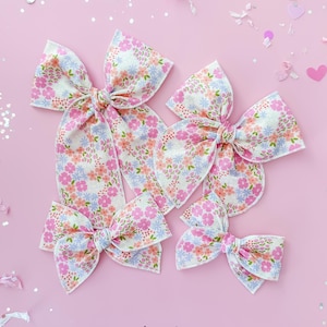 May include: Five fabric hair bows in various sizes, arranged on a pink background. The bows feature a floral pattern with pink, blue, and peach flowers on a white background. The bows are tied in the center with long, flowing tails.
