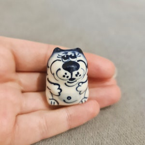 Op de afbeelding: Een klein keramiek beeldje van een witte kat met blauwe details. De kat zit met zijn poten ingetrokken en heeft een cartoonachtige uitdrukking op zijn gezicht.