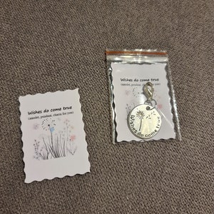 May include: A silver-colored charm with the words "wishes do come true" and a dandelion design, packaged with a card that has the same text and floral illustrations. The charm is in a clear plastic bag.
