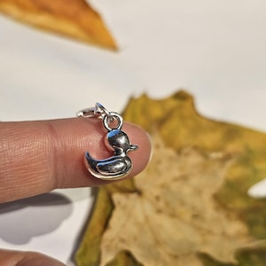 Può includere: Un piccolo ciondolo a forma di anatra color argento con un design dettagliato. Il ciondolo è sospeso da un piccolo anello. Il ciondolo a forma di anatra è tenuto da un dito, con uno sfondo sfocato di una foglia.