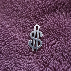 May include: A silver-colored dollar sign charm, displayed against a textured, deep purple background. The charm is a stylized representation of the dollar symbol, commonly used to represent wealth and currency.