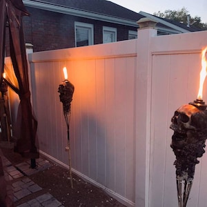 May include: Outdoor scene featuring skull-shaped tiki torches with flames. The torches are mounted on tall poles and are positioned along a white fence. The flames are bright orange against the dusk sky. A dark brown canopy is visible on the left.