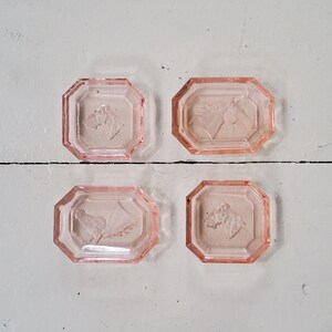 Könnte beinhalten: Vier rosafarbene Glasuntersetzer mit achteckigen Kanten. Jeder Untersetzer hat ein anderes geprägtes Design: einen Pferdekopf, einen Vogel, ein Blumenarrangement und einen Baum. Die Untersetzer sind auf einer weißen Holzoberfläche angeordnet.