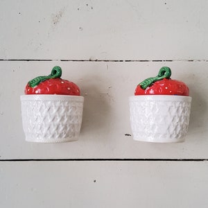 Peut inclure: Deux contenants en céramique blanche avec des couvercles en forme de fraise rouge et verte. Les contenants ont un motif tissé.