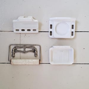 May include: Four vintage bathroom fixtures are displayed against a white wooden background. The items include two white ceramic soap dishes, a white ceramic toilet paper holder, and a metal and ceramic toilet paper holder.