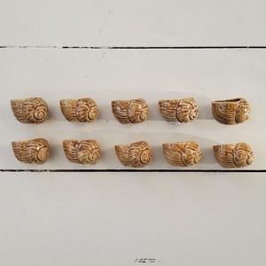 Op de afbeelding: Tien kleine, keramische, slakkenvormige bekers in bruine en beige tinten. De bekers zijn gerangschikt in twee rijen van vijf op een wit houten oppervlak.