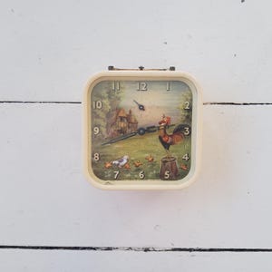 May include: A vintage cream-colored Smith Alarm clock with a square face. The clock face features a pastoral scene with a rooster, chicks, and a cottage. The clock hands are black, and the numbers are in a classic font.